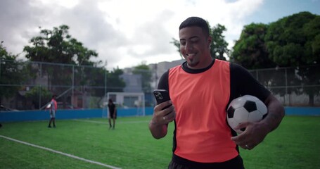 Smiling Latin Hispanic man of African descent checking smartphone while holding soccer ball on green field during friendly match break, digital connection, modern sport lifestyle - Powered by Adobe