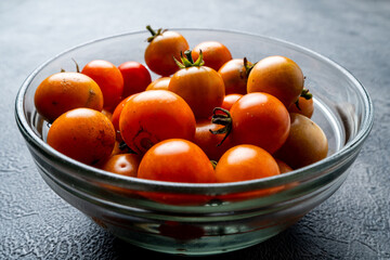 A close-up shot of a clear glass bowl filled with fresh, ripe cherry tomatoes. The bowl rests on a dark, textured slate background. Healthy food ingredient.
