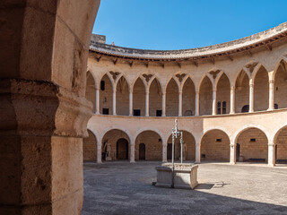 Obraz premium Castell de Bellver. Palma de Mallorca, historic circular courtyard features a stone fountain in the center, surrounded by arched colonnades with a clear blue sky overhead, Majorca