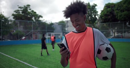 Smiling Latin Hispanic man of African descent standing on soccer field holding ball, using smartphone and presenting green screen display to camera, youth lifestyle, technology, social connection - Powered by Adobe