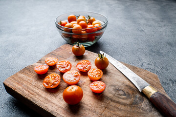 Fresh cherry tomatoes (whole and sliced) on a rustic wooden cutting board with a knife. A bowl of tomatoes is on the dark textured background. Cooking concept.
