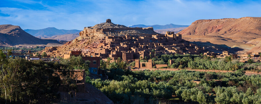 Ancient clay fortress atop desert hill with lush greenery and distant rugged mountains. Ait Benhaddou. Morocco