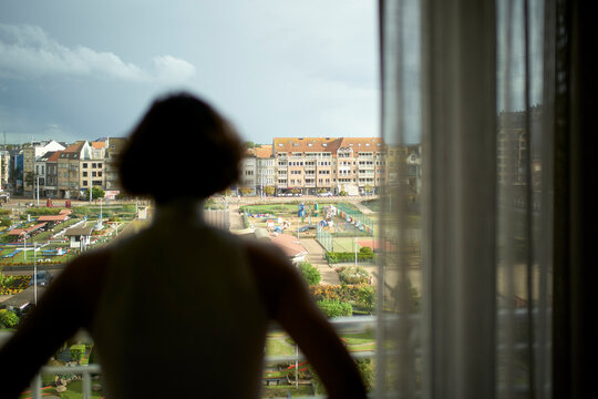 Silhouette of a woman looking out through sheer curtains at a cityscape under a cloudy sky. Blankenberge, West Flanders, Belgium