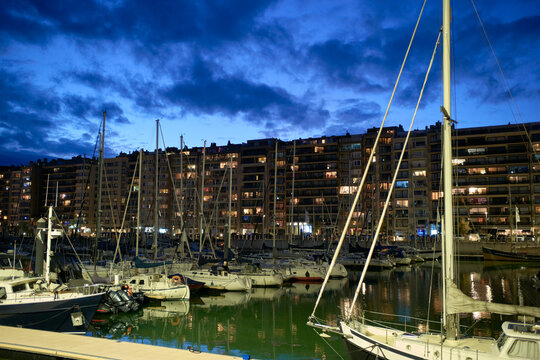Boats docked at a marina with city lights reflecting on the water at twilight. Blankenberge, West Flanders, Belgium