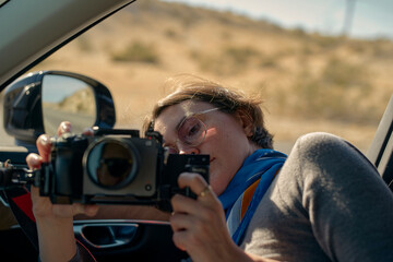 Woman in car adjusting camera, wearing glasses and scarf against warm desert background. California, USA