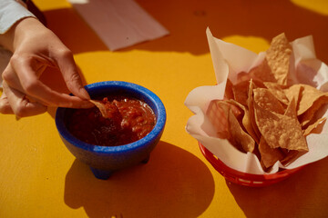 Hand dipping corn chip into salsa bowl on bright orange table next to basket of chips. California, USA