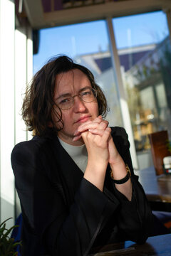 Young woman with glasses sitting in sunlight by a window in a cafe, hands clasped and relaxed. Blankenberge, West Flanders, Belgium