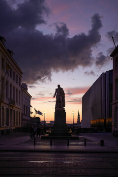 A statue stands silhouetted against a dramatic sunset sky between two buildings in an urban setting. Blankenberge, West Flanders, Belgium