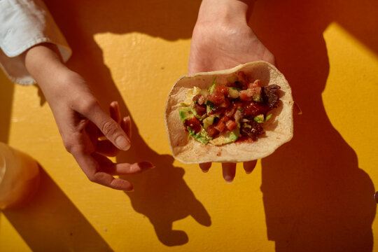 Hands holding a tortilla with salsa and guacamole, on a bright yellow table. California, USA