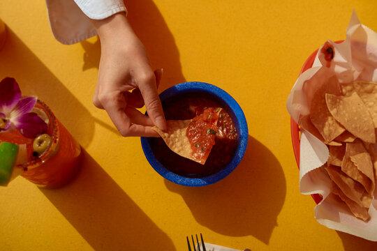 Hand dipping a tortilla chip into salsa with chips, drink, and bright yellow background. California, USA