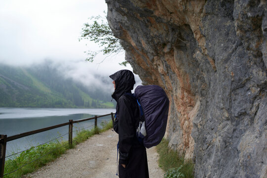 Person in raincoat with backpack on mountain path beside misty lake and rocky cliff. Salzkammergut, Salt Domain, Dachstein Mountains, Austria - Powered by Adobe
