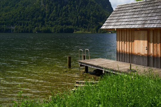 A serene lakeside view with a wooden dock and a rustic cabin surrounded by lush greenery. Salzkammergut, Salt Domain, Dachstein Mountains, Austria