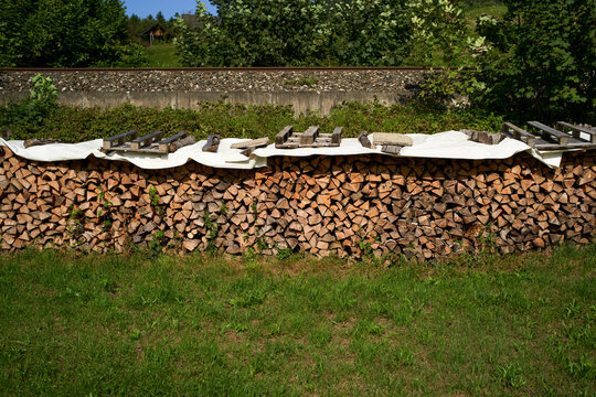 A large stack of firewood neatly piled on grass with a white cover and wooden planks on top. Salzkammergut, Salt Domain, Dachstein Mountains, Austria