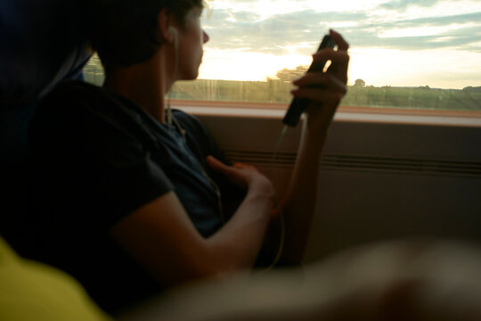 Young man on a train wearing earphones, looking out window at sunset holding phone. Munich, Germany