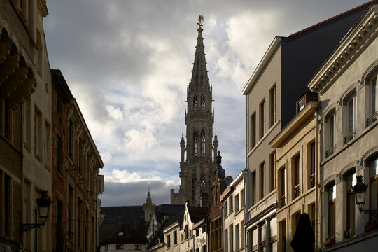 A narrow European street with historic buildings and a tall, ornate church tower under cloudy skies. Brussels, Belgium