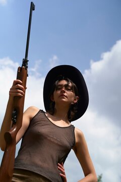 Young woman in a mesh top and cowboy hat holds a rifle on a cloudy day.