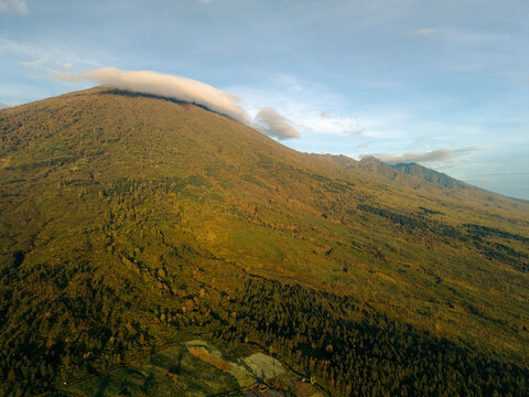 Aerial view of a vast mountain landscape bathed in golden sunlight with a cloud-topped peak. Lombok, Senaru, Indonesia