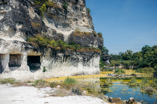 Rocky cliffside with caves, adjacent to a small pond surrounded by greenery under a blue sky. Madura, East Java, Indonesia