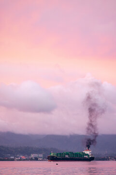 Cargo ship with black smoke sails under a pink sky at sunrise over calm waters. Ambon, Maluku, Indonesia