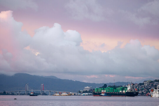 Cargo ship with green containers at sea under colorful sunset clouds near a coastal city. Ambon, Maluku, Indonesia
