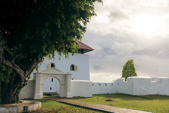 A white fortress with an archway under a large tree, surrounded by greenery and a cloudy sky. Ambon, Maluku, Indonesia