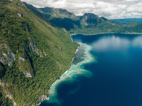 Aerial view of lush green mountains meeting a vibrant blue ocean with a coral reef fringe. Saleman Village, Seram Island, Indonesia