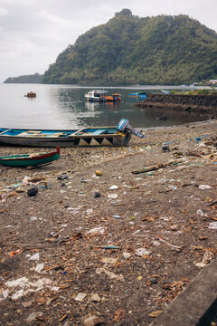 A plastic-polluted beach with boats and a lush green mountain by the sea. Banda Neira, Banda, Central Maluku, Indonesia