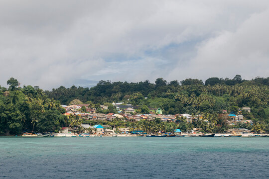 Coastal village with houses, lush greenery, and turquoise sea under a cloudy sky. Pulau Rhun or Rhun Island, Banda, Central Maluku Regency, Indonesia