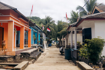 Colorful houses line a narrow street with palm trees and flags on a cloudy day. Pulau Rhun or Rhun Island, Banda, Central Maluku Regency, Indonesia