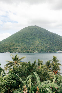 Lush green mountain rises over a serene lake with palm trees and cloudy sky. Banda Neira, Banda, Central Maluku Regency, Indonesia