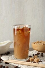 Tasty iced coffee with milk in glass, beans and sugar cubes on wooden table against grey background, closeup