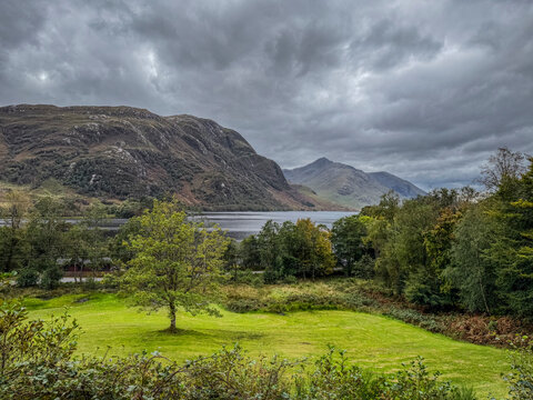 A scenic landscape with lush green grass, trees, mountains, and a cloudy sky over a lake. Glenfinnan, Loch Shield, Scotland