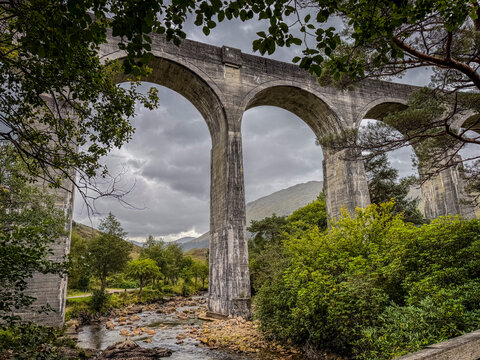 Majestic stone viaduct arches over lush green valley with a serene flowing river below. Glenfinnan Viaduct, Scotland