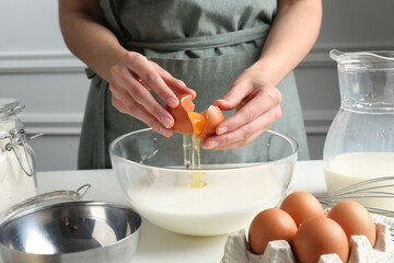 Making batter (liquid dough). Woman adding eggs into bowl at white table indoors, closeup