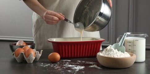 Woman pouring liquid dough from bowl into baking dish at grey table in kitchen, closeup