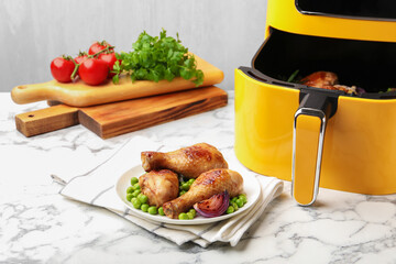 Air fryer with grilled chicken drumsticks and ingredients on white marble table, closeup