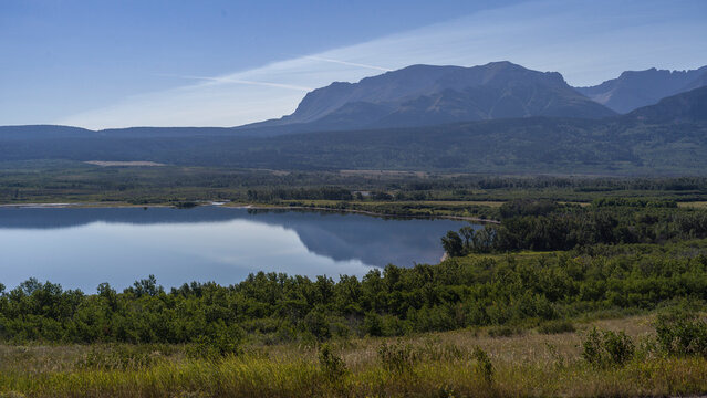 Serene lake surrounded by lush greenery and distant mountains under a clear blue sky. Waterton Park, Alberta, Canada - Powered by Adobe