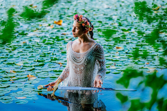 Woman in white lace dress holds a sword while standing in a lily-covered pond. Wildcat Lake Park, WA, USA