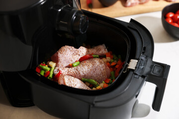 Air fryer with raw chicken drumsticks and vegetables on table, closeup