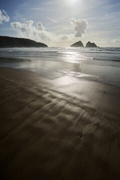 Holywell Bay and beach, Cornwall, UK. Sunlit coastal landscape with wet sand and distant rocky islets under a cloudy sky.