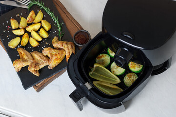 Air fryer with grilled vegetables, chicken wings, potato wedges and spices on white marble countertop, flat lay