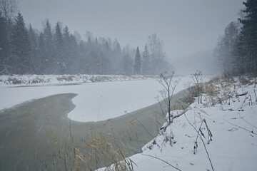 Obraz premium Landscape freezing river and snowfall against the backdrop of a winter forest and snow in cloudy weather