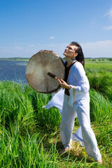Middle-aged female shaman in the white dress drumming outdoors