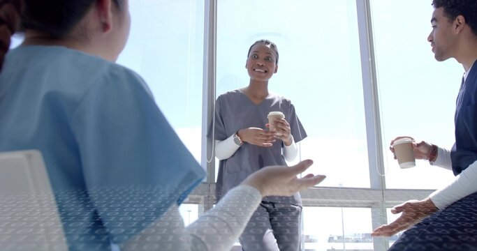Chatting medical team in scrubs holding two paper coffee cups by floor-to-ceiling windows in lounge