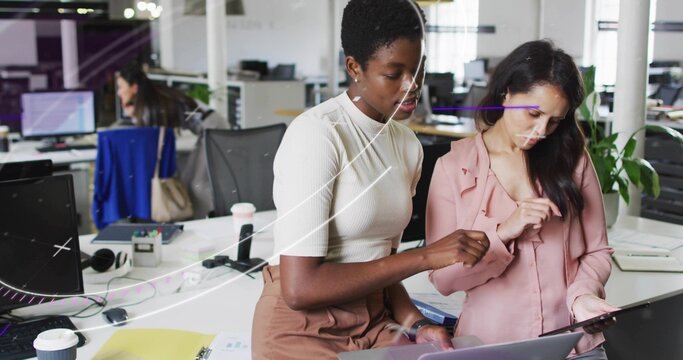 Collaborating coworkers wearing cream top and light pink blouse pointing at laptop in modern office