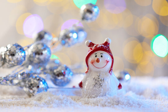 Cute decorative snowman on artificial snow against blurred lights, closeup