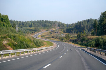 A long highway winds through peaceful countryside, lined with trees on both sides. A white vehicle travels along the road on a clear day, showcasing the natural beauty of the area.