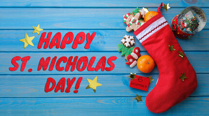Happy St. Nicholas Day greeting card design with red boot full of sweet treats and snow globe on blue wooden background, top view