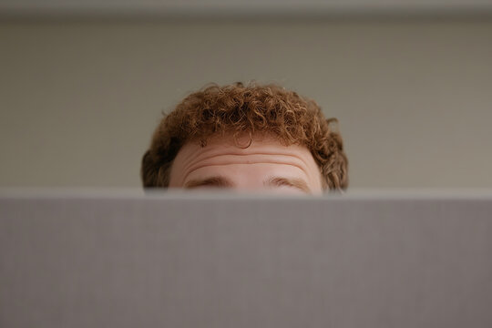 A man with curly brown hair peeks over a gray cubicle wall, eyes peering forward. The neutral background blurs the line between focus and anonymity in the modern workspace.