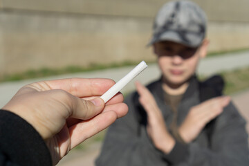 hand offering a white cigarette to a young boy in a cap who is blurred and making a clear no, stop gesture with his arms, concept of youth health, antismoking campaign, education